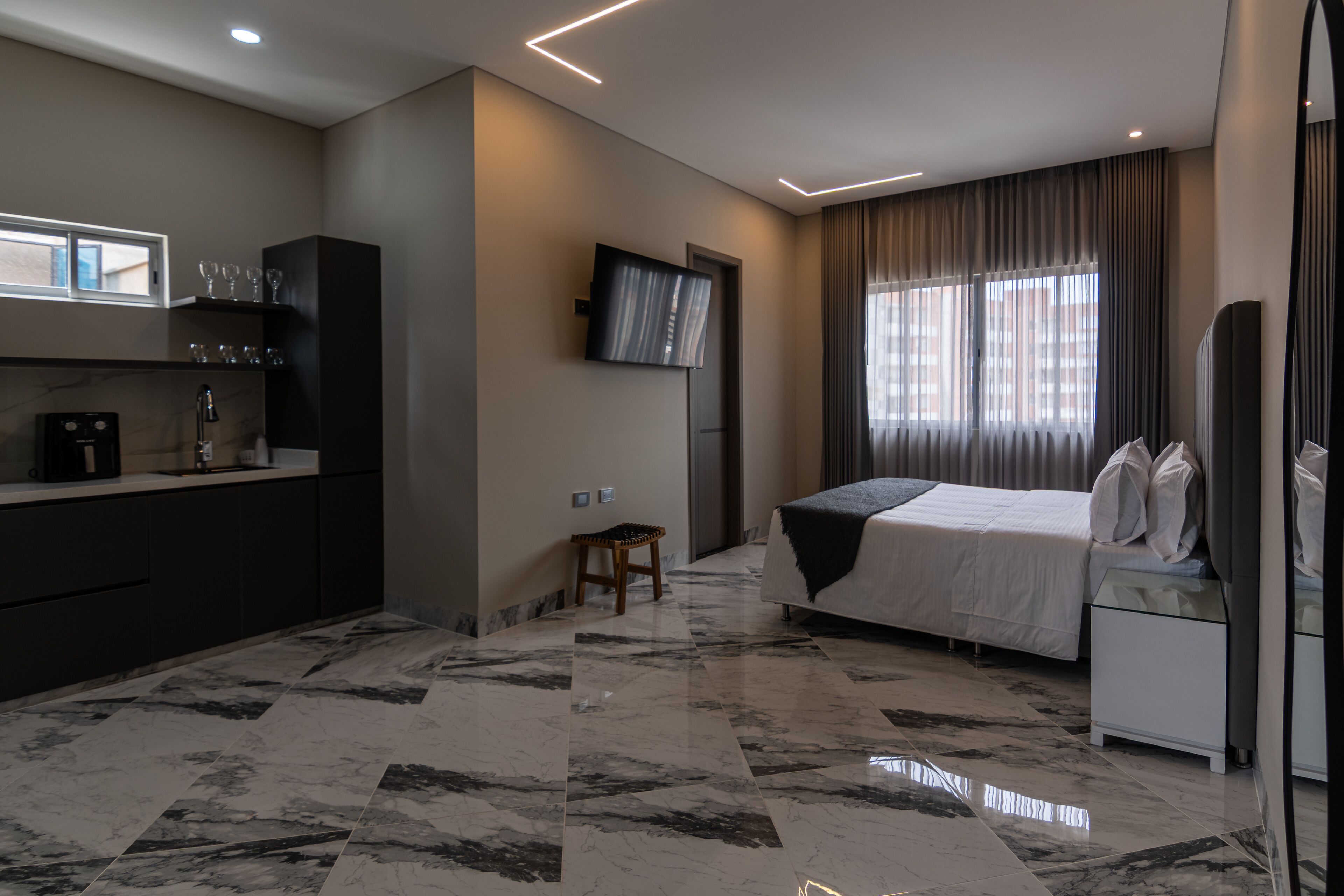Ein modernes Hotelzimmer mit Bett, Wandfernseher, einem kleinen Hocker und einem Schreibtisch mit Kaffeemaschine.