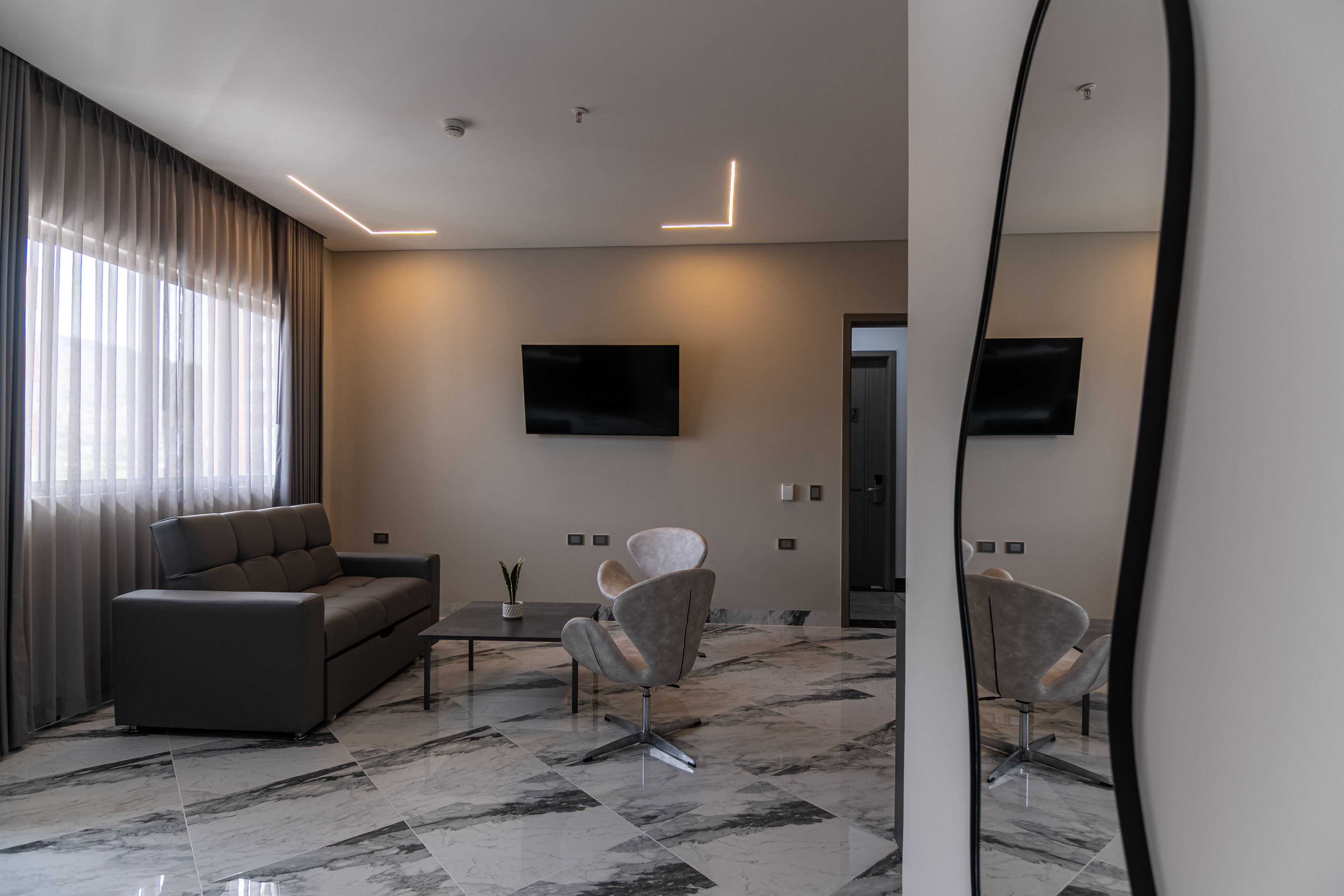 A modern living room with a sofa, armchair, and coffee table, a flat-screen TV mounted on the wall, and a large mirror reflecting the room.