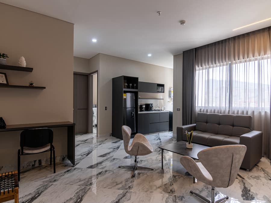 Una sala moderna con un sofá, dos sillones, una mesa de centro y una zona de cocina con armarios y electrodomésticos.