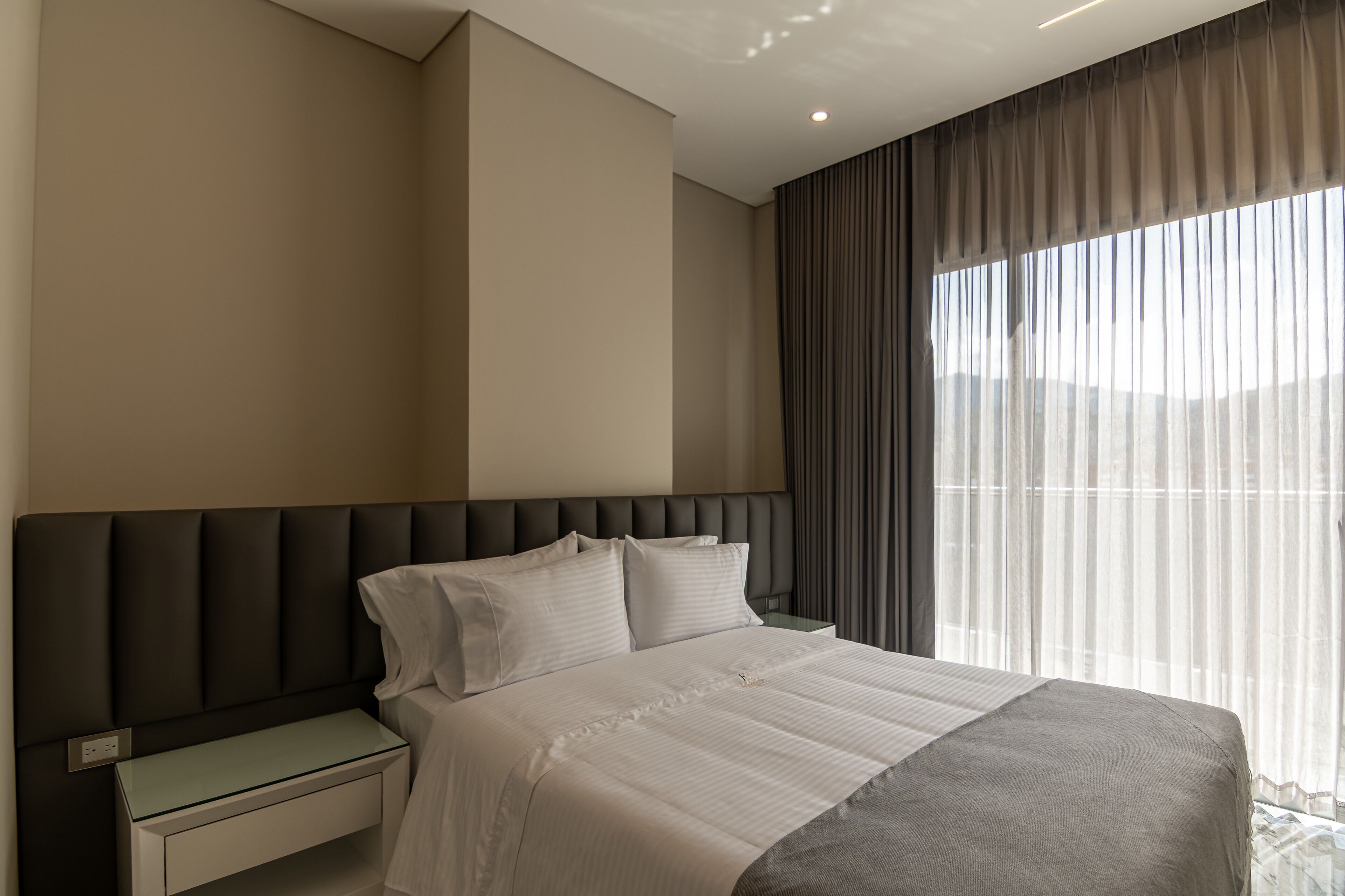 A modern hotel room with a large bed, a nightstand, and a view through sheer curtains.