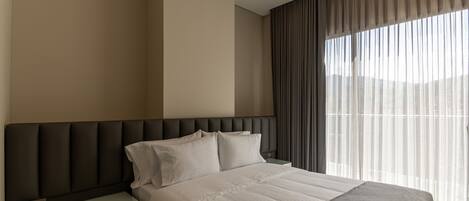 A modern hotel room with a large bed, a nightstand, and a view through sheer curtains.