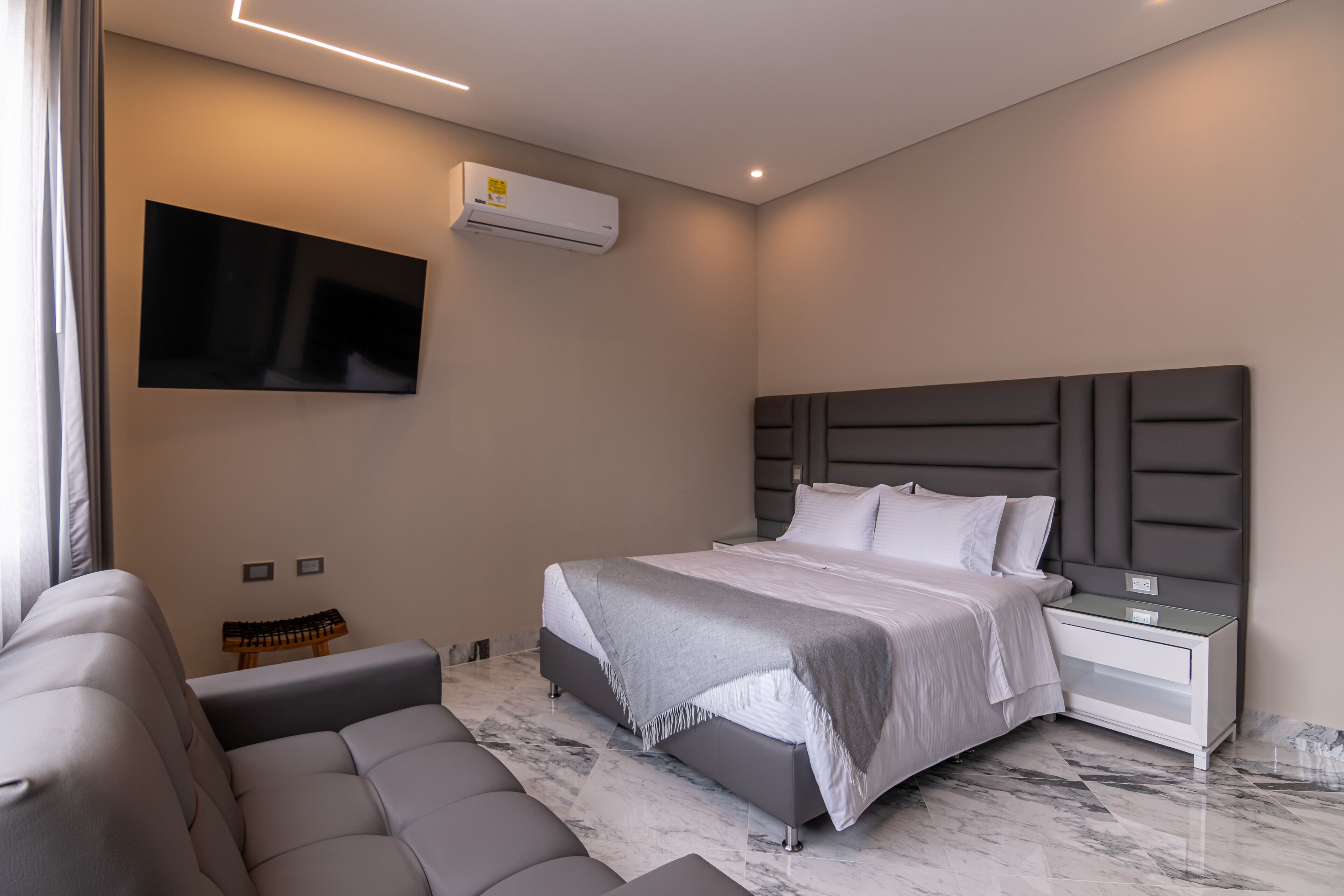 A modern bedroom with a bed, a sofa, a TV, and an air conditioning unit.