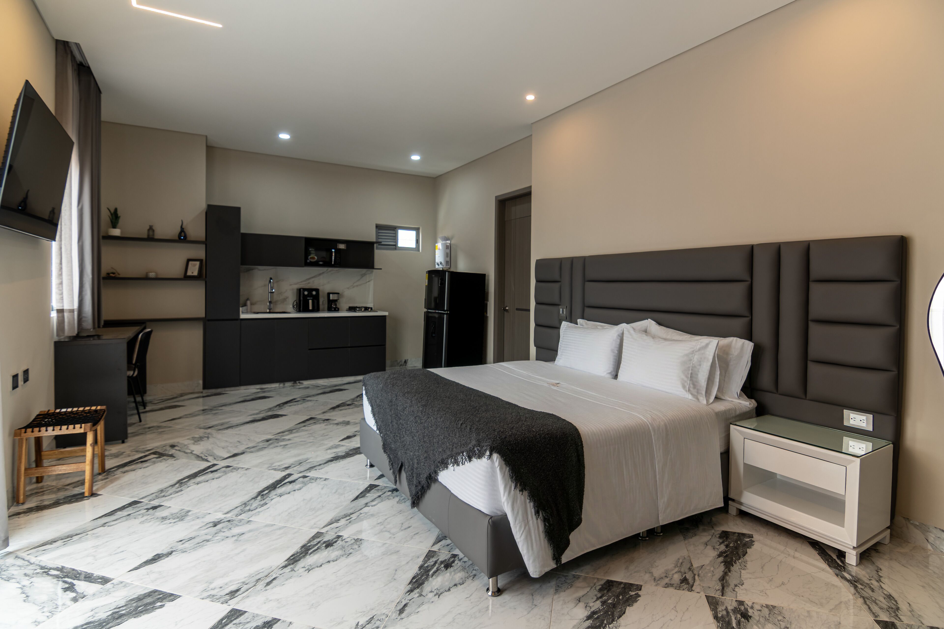 A modern hotel room with a large bed, a bedside table, a built-in desk, and a kitchen area.