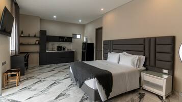 A modern hotel room with a large bed, a bedside table, a built-in desk, and a kitchen area.
