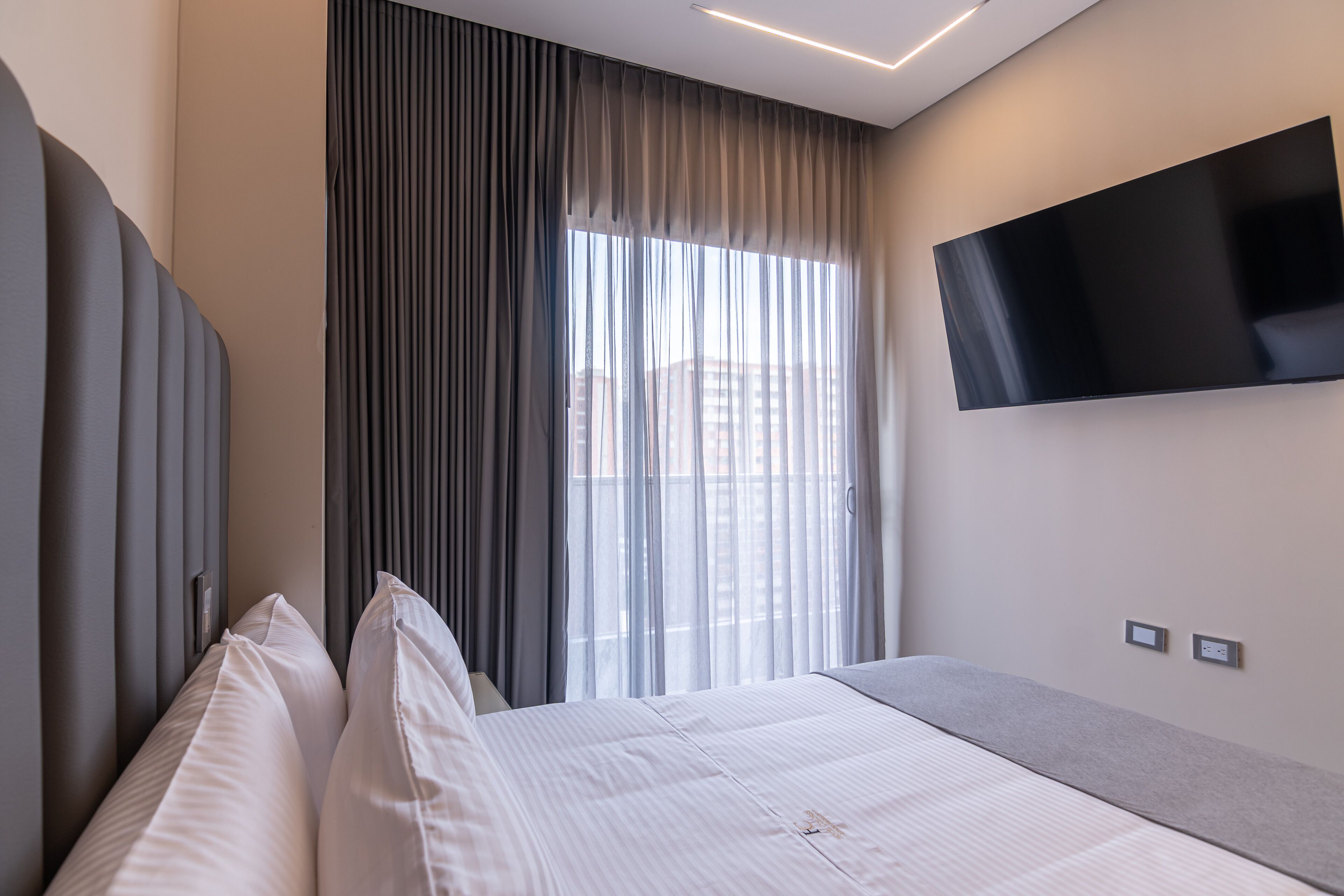 A modern hotel room with a large bed, a flat-screen TV mounted on the wall, and a window with sheer curtains.