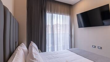 Ein modernes Hotelzimmer mit einem großen Bett, einem flachbildfernseher an der Wand und einem Fenster mit durchsichtigen Vorhängen.