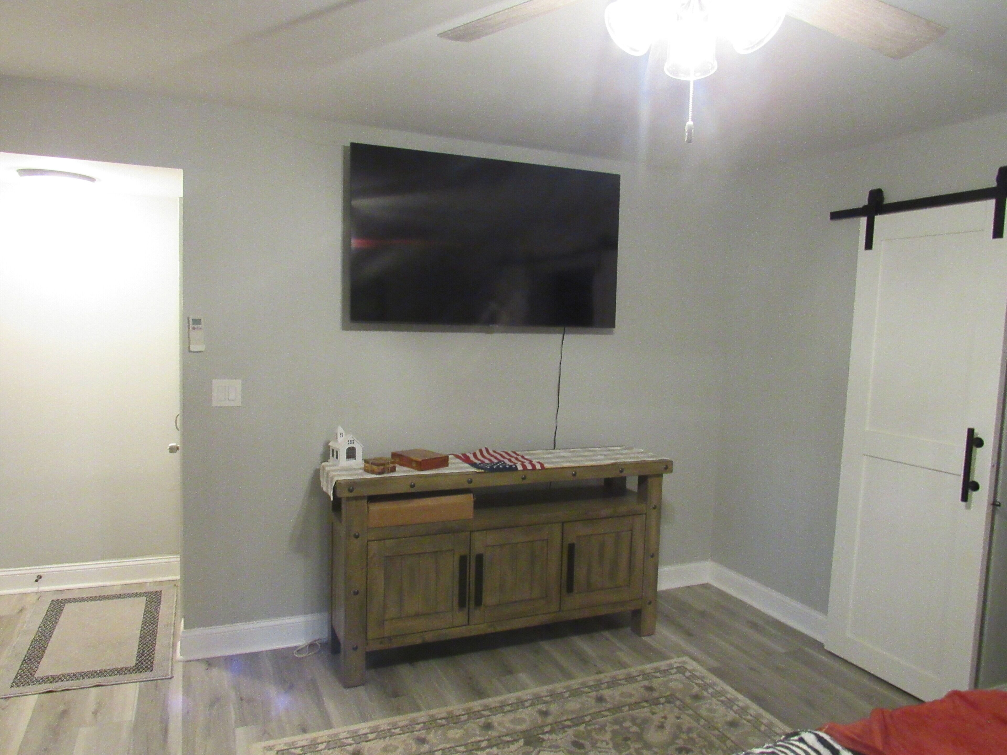 A room with a ceiling fan, a television mounted on the wall, a wooden cabinet with drawers, a patterned rug, and a door with a black handle.