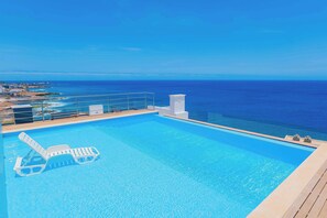 A swimming pool with lounge chairs, a view of the ocean, and a clear sky. - Coastal Waves - Relaxing 3BR Apartment with pool & Ocean Views (Praia)