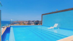 Une piscine d’un bleu éclatant, dotée d’une terrasse en bois et d’une chaise longue, avec vue sur une ville côtière.