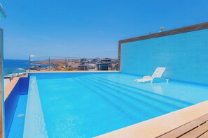 A clear blue swimming pool with a wooden deck and a lounge chair, overlooking a coastal town.