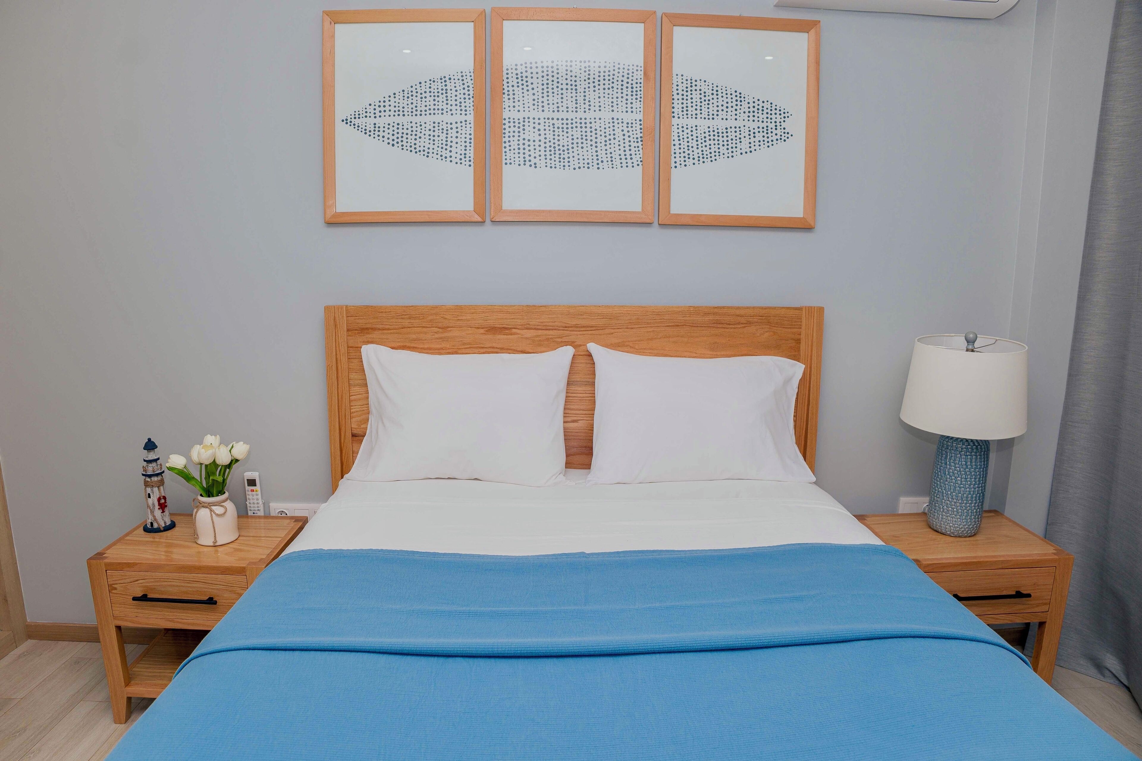 Una cama bien hecha con una colcha azul, dos mesitas de noche con lámparas, un jarrón con flores y tres cuadros en la pared.
