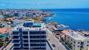A modern building with a rooftop pool, located near a coastal area with a view of the sea. - Coastal Waves - Relaxing 3BR Apartment with pool & Ocean Views (Praia)