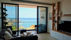 A modern living room with a large TV, a sofa, and a balcony with a view of the sea.
