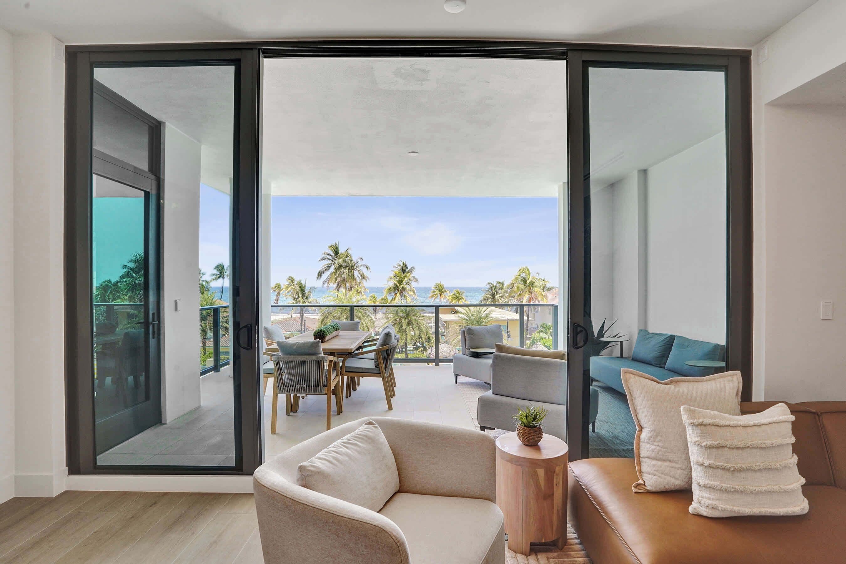 A modern living room with a large sliding glass door leading to a balcony with a view of the ocean and palm trees.