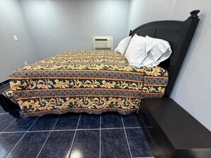 A bed with a patterned bedspread, a white pillow, and a black headboard in a room with tiled flooring.