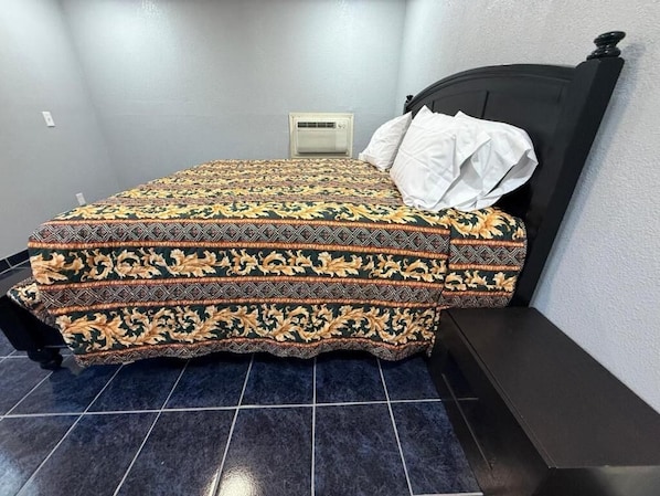 A bed with a patterned bedspread, a white pillow, and a black headboard in a room with tiled flooring.
