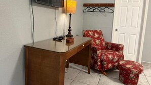 Een kamer met een houten bureau, een stoel, een aan de muur bevestigde televisie, een lamp en een kapstok.