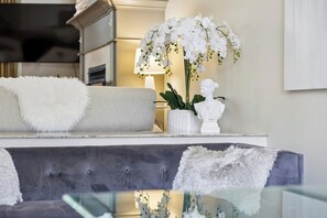 Ein modernes Wohnzimmer mit einem grauen, gesteppten Sofa, weißen, flauschigen Zierkissen, einer weißen Orchideenpflanze und einer weißen Büste auf einem weißen Marmortisch.