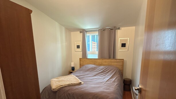 Un dormitorio con una cama de madera, una ventana con cortinas, una mesita de noche con lámpara y una puerta.