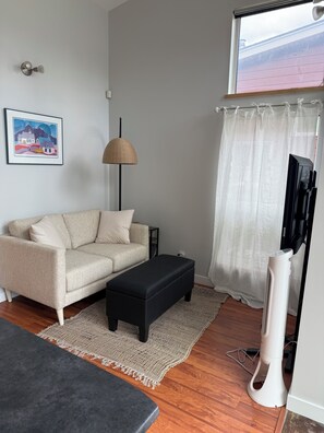 A living room with a sofa, an ottoman, a floor lamp, a TV, and a fan.
