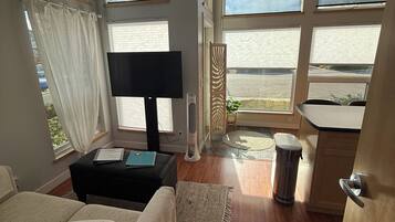 A room with a sofa, a TV, a small table, a fan, and a view of the outdoors through large windows.