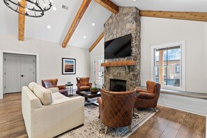A spacious living room with a stone fireplace, leather armchairs, a sofa, and a flat-screen TV mounted above the fireplace.