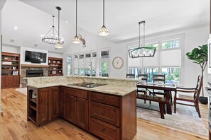 A spacious kitchen with a large island, wooden cabinets, and a stone fireplace. There is a dining area with a table and chairs, and a large window providing a view of the outdoors. - McLemore Resort - 84 Rushing Water Trail - Forest and Pond View (Rising Fawn)