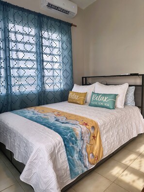 Ein Bett mit einem Strand-Themen-Bettwäschebezug, ein Fenster mit blauen Vorhängen und eine Wand-Klimaanlage.