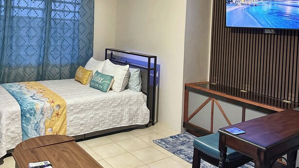 A hotel room with a bed, a TV mounted on a wooden panel, a ceiling fan, an air conditioning unit, and a small wooden table. - Peaceful Studio Retreat Near Shoal Bay Beach - AC, Wi-Fi, Concierge Support (Shoal Bay Corner)