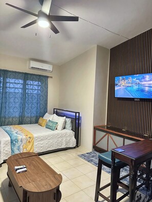 A hotel room with a bed, a TV mounted on a wooden panel, a ceiling fan, an air conditioning unit, and a small wooden table. - Peaceful Studio Retreat Near Shoal Bay Beach - AC, Wi-Fi, Concierge Support (Shoal Bay Corner)