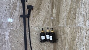 A shower with a handheld showerhead, a handheld scrubber, and three bottles of shampoo, conditioner, and body wash on a wall-mounted rack. - Peaceful Studio Retreat Near Shoal Bay Beach - AC, Wi-Fi, Concierge Support (Shoal Bay Corner)