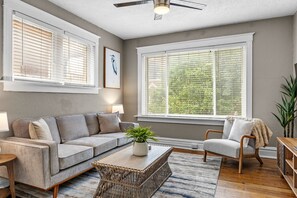 A living room with a sofa, armchair, coffee table, and a large window with blinds. - The Cove | A St. Louis Retreat by StayLage (Saint Louis)