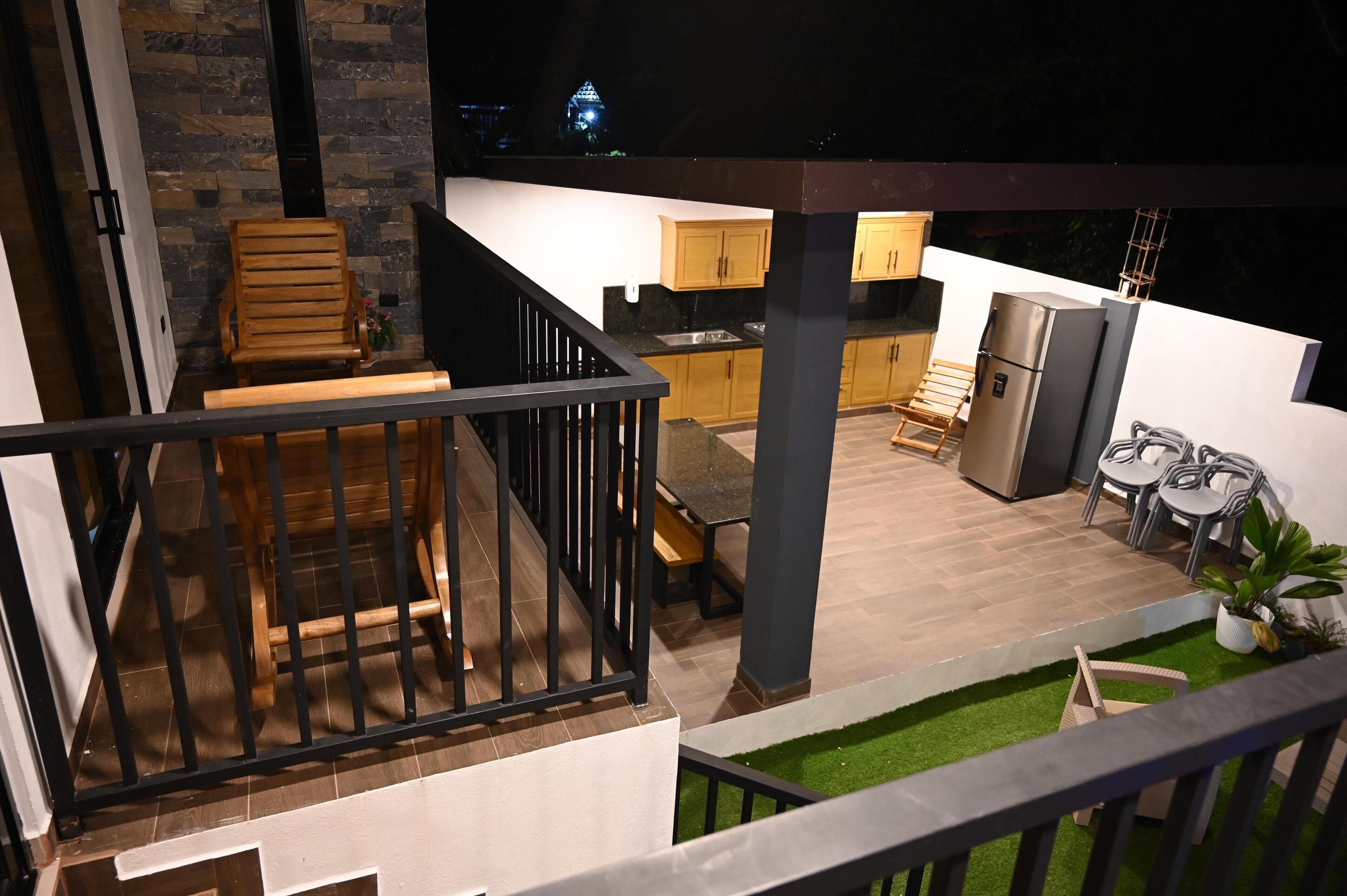 A rooftop patio with a wooden deck, a seating area with chairs, a small kitchen with a refrigerator, and a view of a distant building.