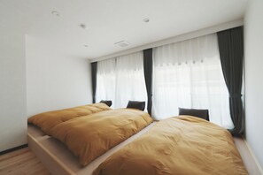 A bedroom with two beds, a window with sheer curtains, and a wooden floor.