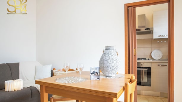 Un coin repas avec une table en bois, deux verres à vin, un vase décoratif et une cuisine visible à travers une porte ouverte.