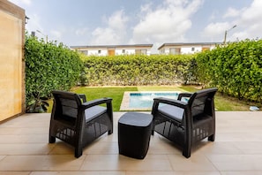 A patio with two wicker chairs and a small table, a swimming pool, and a neatly trimmed hedge. - Vista Cana private Jacuzzi + Golf + Beach + Pool (Punta Cana)