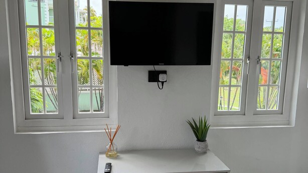 A white room with a flat-screen TV mounted above a white dresser with drawers, two windows with open shutters, and a small potted plant on the dresser. - Cap Estate 3BR Townhome, Private Plunge Pool, Golf (Gros Islet)