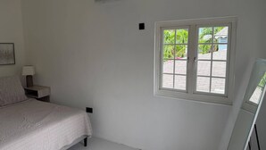 A bedroom with a bed, a bedside table with a lamp, a window with a view of greenery, and an air conditioning unit. - Cap Estate 3BR Townhome, Private Plunge Pool, Golf (Gros Islet)
