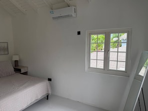 Ein Schlafzimmer mit einem Bett, einem Nachttisch mit Lampe, einem Fenster mit Blick ins Grüne und einer Klimaanlage.