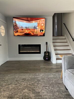 Un salon avec un téléviseur à écran plat fixé au mur, une cheminée en dessous, une guitare appuyée contre le mur et un escalier sur la droite.