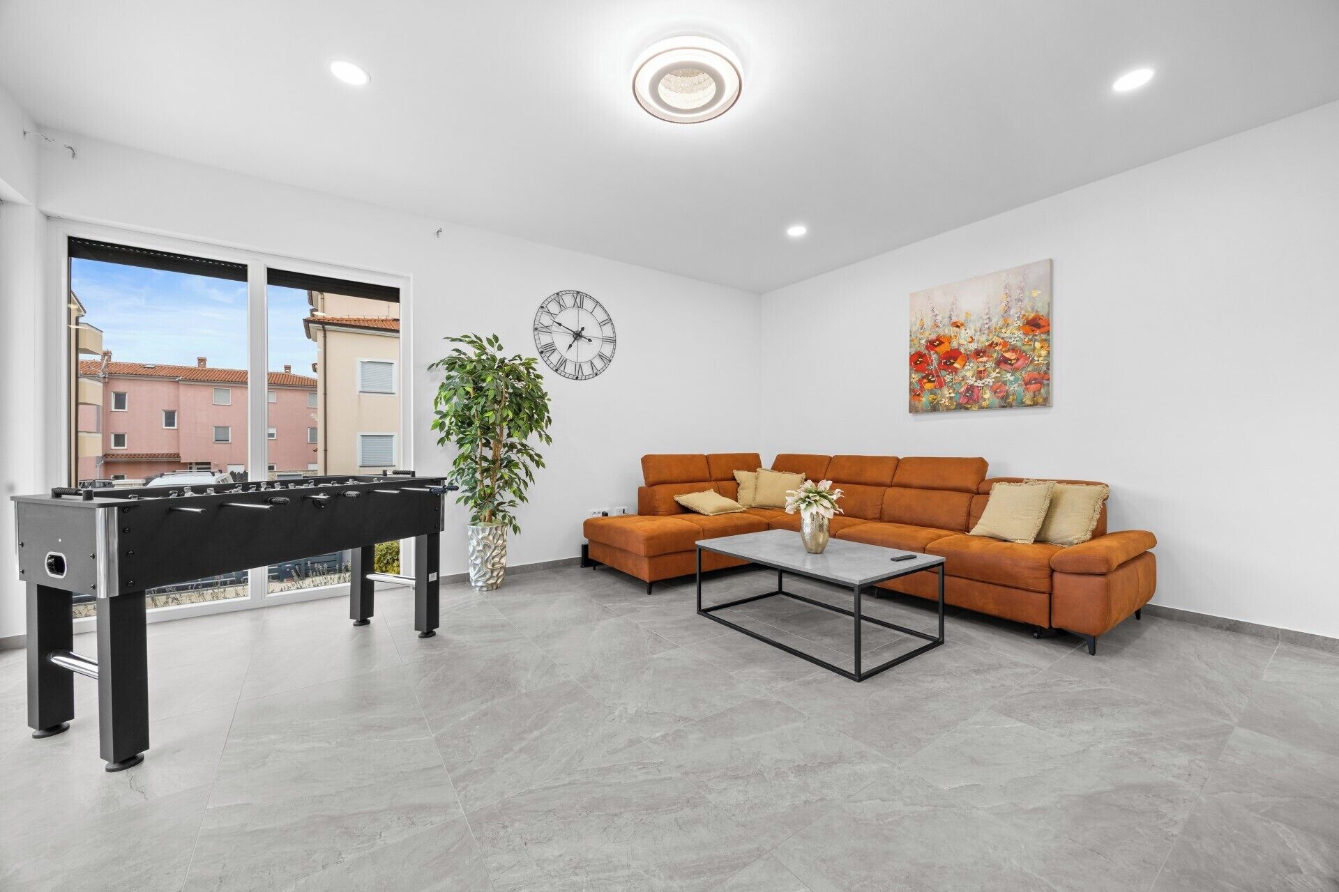 A modern living room with a foosball table, a sofa, a coffee table, a clock, and a painting.