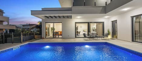 Une maison moderne dotée d’une piscine, d’un coin repas et d’une cuisine visible à travers des portes vitrées.