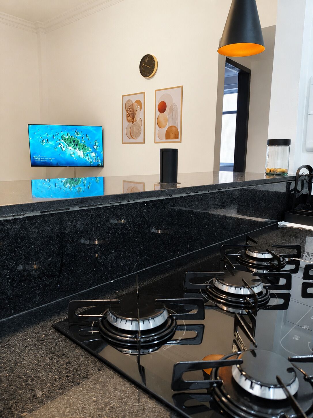 Uma cozinha com bancada preta, quatro bocas de gás, coifa preta, televisão fixada na parede e quadros decorativos.