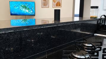 Uma cozinha com bancada preta, quatro bocas de gás, coifa preta, televisão fixada na parede e quadros decorativos.