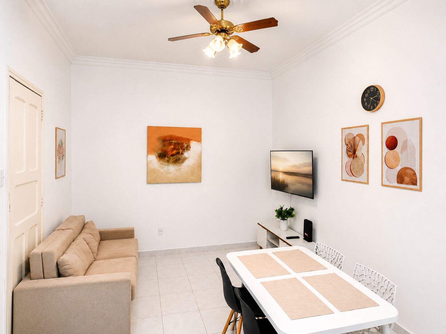 Uma sala de estar moderna com um sofá bege, uma mesa de jantar com quatro cadeiras, um ventilador de teto, uma TV de tela plana e obras de arte nas paredes.