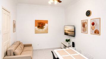 Uma sala de estar moderna com um sofá bege, uma mesa de jantar com quatro cadeiras, um ventilador de teto, uma TV de tela plana e obras de arte nas paredes.