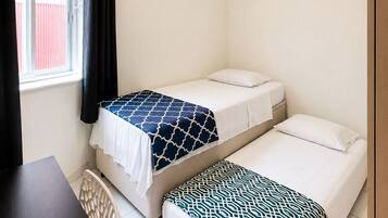 Quarto com duas camas de solteiro, um ventilador de teto, janela com cortinas e uma escrivaninha.