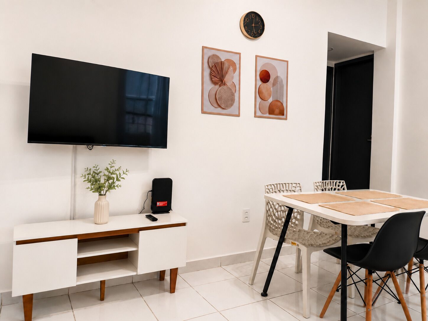 Uma sala de estar moderna com uma TV de tela plana, uma escrivaninha branca com uma planta, um alto-falante preto e um espaço de jantar com mesa e cadeiras.