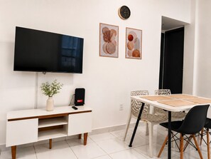 Uma sala de estar moderna com uma TV de tela plana, uma escrivaninha branca com uma planta, um alto-falante preto e um espaço de jantar com mesa e cadeiras.