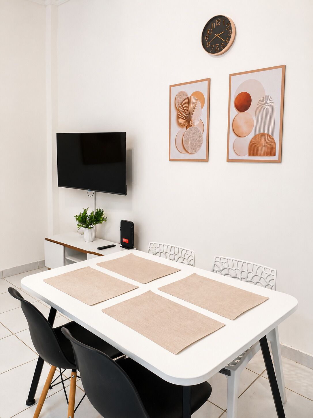 A modern dining area with a white table, black chairs, a flat-screen TV, a clock, and two framed artworks.